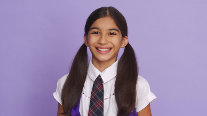 Funny positive indian kid primary school girl with ponytails wearing uniform laughing grimacing looking at camera on violet background. Happy latin child student close up headshot portrait. Royalty-Free Stock Footage #1058989079