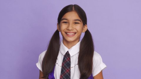 Funny positive indian kid primary school girl with ponytails wearing uniform laughing grimacing looking at camera on violet background. Happy latin child student close up headshot portrait.