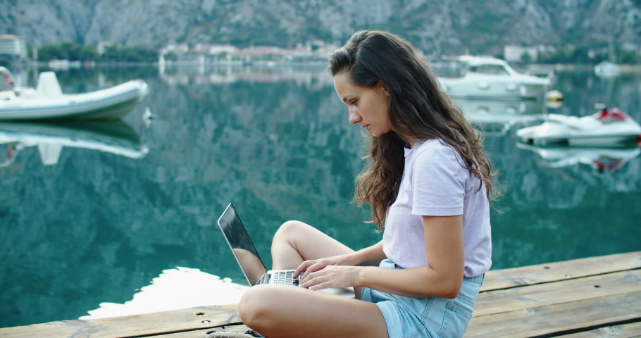 A young woman is sitting on the pier and working on a laptop. Finishes work, closes laptop and rests. Breathes fresh sea air. Freelance. Sea view. The concept of favorite work. High quality 4k footage | Shutterstock HD Video #1058990396