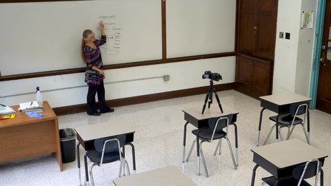 Wide angle view of smiling female english grammar teacher, writing on whiteboard in a school classroom with empty chairs. High quality footage