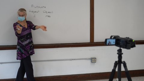 Female english grammar teacher writing on whiteboard in a school classroom wearing a medical face mask for remote hybrid learning in front of video camera.