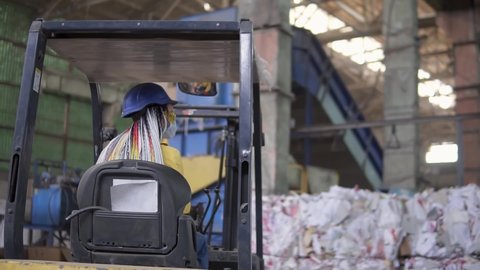 Unloading of waste paper in a warehouse electric car. Female worker in hard hat and yellow jacket sitting in machine. Huge stocks of pressed carton. Woman driver sits from the back and pointing on