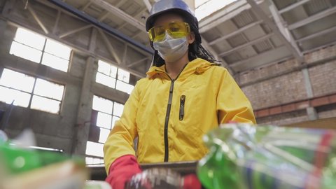 Woman-volunteer in yellow jacket and transparent protecting glasses, hard hat and mask sorting used plastic bottles at recycling plant. Separate bottles on the line, removing tops and squeeze them