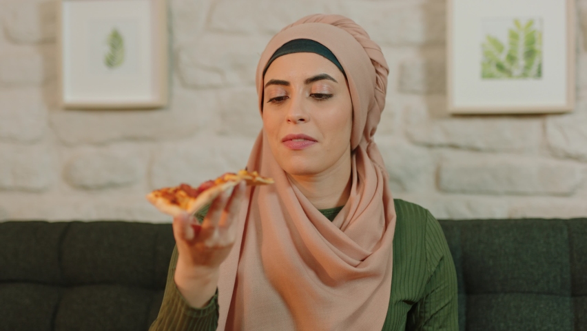 A cheerful young woman in a turban is sitting on an armchair at home and eating pizza.