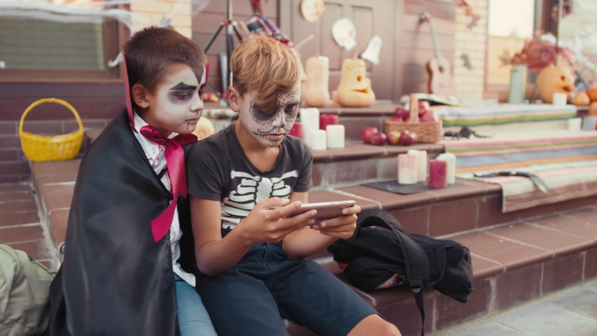Medium shot of boys wearing scary makeup and halloween costumes sitting on porch of house and playing on mobile phone