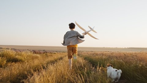 Happy boy walk Wheat Field holding airplane in his hand, his dog Jack Russell Terrier runs on summer meadow against of sunset of summer day towards bright sun slow motion. Childhood plays lifestyle