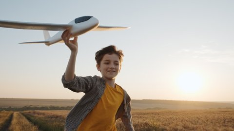 Happy boy runs across field of golden wheat holding airplane in his hand against of sunset of summer day towards bright sun slow motion. Child plays lifestyle. Travel. Childhood. Lifestyle. Agro. Farm