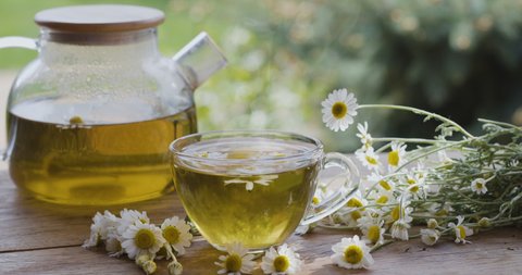 Chamomile tea in a glass teapot and cup, the camera moves around. The action takes place on a summer rural terrace with playing sunbeams. Nearby are the flowers of a pharmaceutical chamomile.    