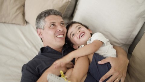 Joyful dad cuddling and tickling laughing little son on couch in living room. Boy and his father roping and playing together. Family fun time concept