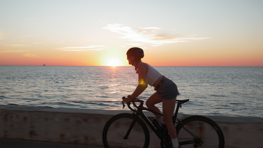 Young stylish brunette woman rides bike on coastline at sunrise. Brunette girl in helmet glasses and stylish clothes is pedaling on bicycle at sunset Royalty-Free Stock Footage #1059004433