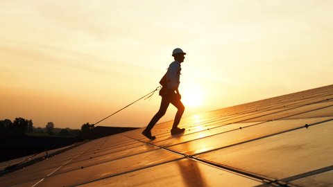 Maintenance assistance technical worker in uniform is checking an operation and efficiency performance of photovoltaic solar panels on roof at sunset.