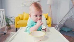 infant boy sitting in baby chair and sucking spoon near bowl