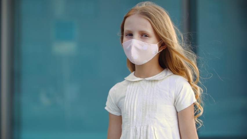 Serious young girl with long golden blonde curls wears white dress and pink protective face mask, stands posing outdoors in front of camera, looking upset with resentment, pandemic epidemic virus Royalty-Free Stock Footage #1059016106