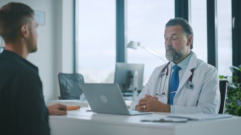 Family Doctor is Delivering Great News About Male Patient's Medical Results During Consultation in a Health Clinic. Physician in White Lab Coat Sitting Behind a Computer in Hospital Office.