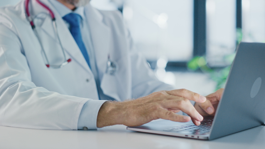 Close Up of a Family Medical Doctor Working on a Laptop Computer in a Health Clinic. Physician in White Lab Coat is Browsing Medical History Behind a Desk in Hospital Office. Royalty-Free Stock Footage #1059016322