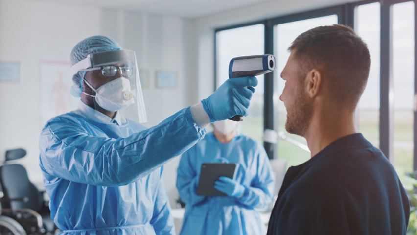 Medical Physician in Safety Gear Measuring Male Patient's Body Temperature with Infrared Thermomether in a Health Clinic. Doctor Uses Touch-free Technology to Diagnose Covid-19 Symptoms. | Shutterstock HD Video #1059016361