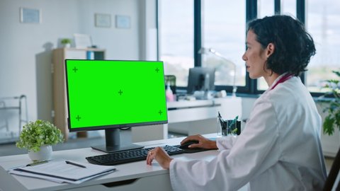 Female Medical Doctor is Working on a Computer with Green Screen Mock Up Display in a Health Clinic. Assistant in White Lab Coat is Reading Medical History Behind a Desk in Hospital Office.