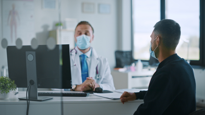Family Doctor in Protective Mask is Reading Medical History of Young Male Patient and Speaking with Him During Consultation in a Health Clinic. Physician in Lab in Front of Computer in Hospital Office | Shutterstock HD Video #1059016538