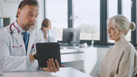 Friendly and Cheerful Family Doctor is Reading Medical History of Senior Female Patient During Consultation in a Health Clinic. Physician Using Tablet Computer in Hospital Office.