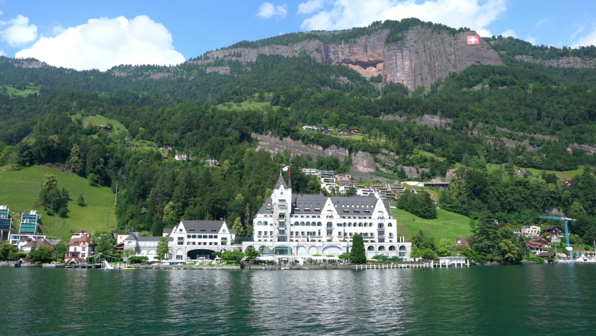 Panorama of Lake Lucerne shoreline with beautiful buildings and mountains with Swiss flag taken from a moving cruise boat in Vitznau Switzerland