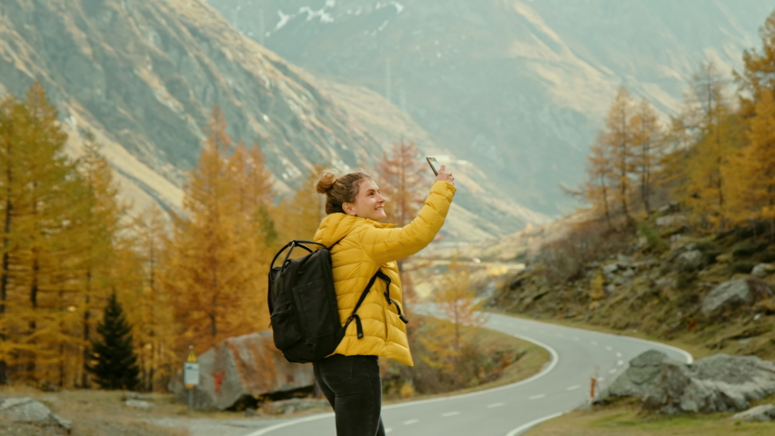 Woman traveller on hike in epic autumn mountains. Happy and excited young woman in outdoor adventure gear outfit walk on beautiful mountain road, spin around, record memories and experiences on phone | Shutterstock HD Video #1059029486