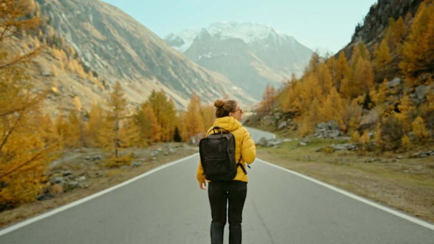 Camera follows young female traveller in yellow puffy jacket and backpack, walk in middle of epic empty road in mountains, surrounded by nature, autumn or fall colours. Travel road trip wanderlust | Shutterstock HD Video #1059029489