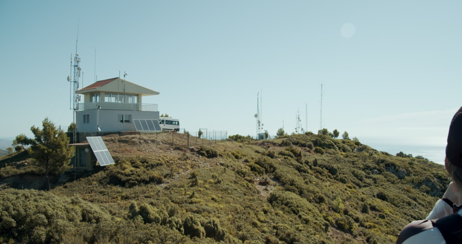 Blonde woman wearing a cap in front of a  mediterranean forest guard post looking at the sea on top of a mountain on a sunny day in a Mediterranean area.