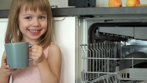 Close up of little girl looks at camera, smiles and laughs, puts cup in dishwasher. Happy child helps with housework. Household and dishwashing concept. Loading dishwasher