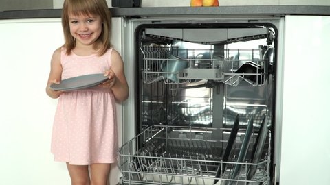 Portrait of little happy girl looks at camera and smiles, puts plate in dishwasher. Child helps with housework. Household and dishwashing concept. Loading dishwasher