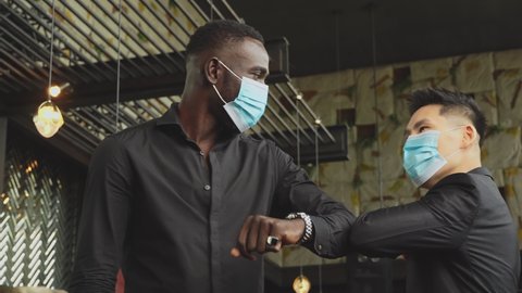 Handheld: African American black man and Asian friend wearing protective face mask elbow bump greeting for social distancing and new normal business etiquette to prevent Coronavirus infection