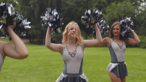 Young cheerful girls with pom poms dancing outdoors. Portrait of cheerleading team in uniform performing cheering dance on green lawn in park