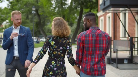 Back view of young woman looking and flirting with other man walking with angry african boyfriend outdoors. Love triangle, betrayal and infidelity concept