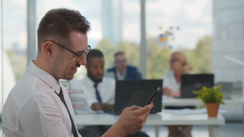 Smiling young man using smartphone working in modern office. Side view of male employee typing message on mobile phone on break at work with colleagues working on background