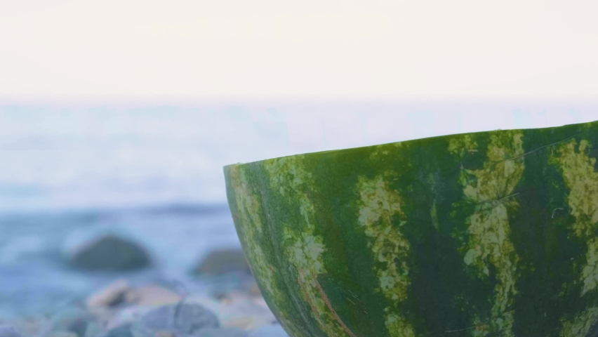 Half Of Ripe Watermelon On The Pebbles In The Beach With Splashing Waves On The Background In Greece. - selective focus shot