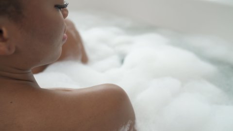 Back view of African American woman taking bubble bath, washing herself with shower sponge. Camera in movement. Spa concept