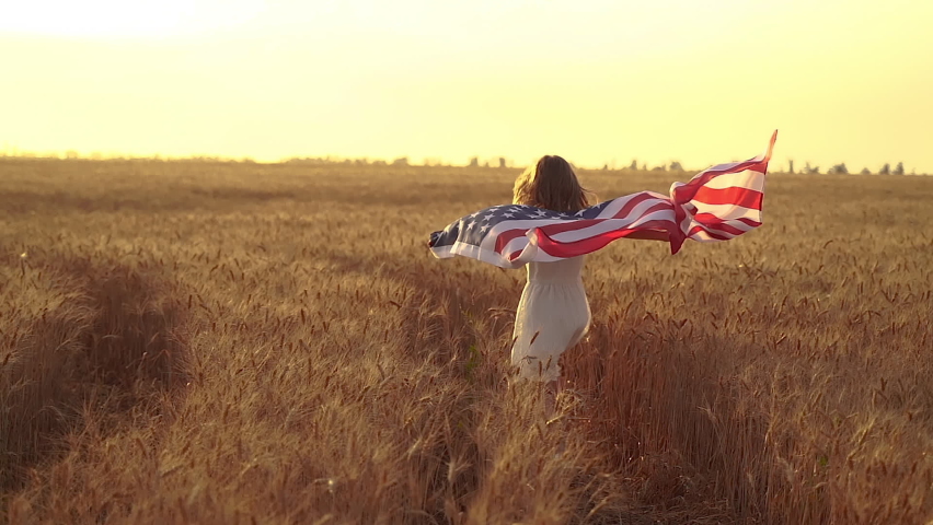 Girl in white dress wearing an American flag while running in a beautiful wheat field on sunset