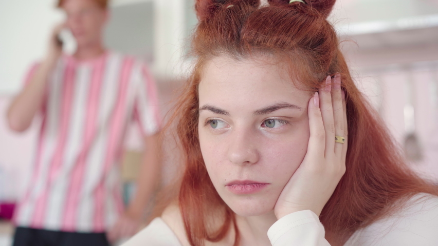 Close-up portrait of gorgeous sad woman holding head with hand. Blurred man talking on the phone at the background. Beautiful redhead Caucasian girlfriend dissatisfied with boyfriend ignorance.