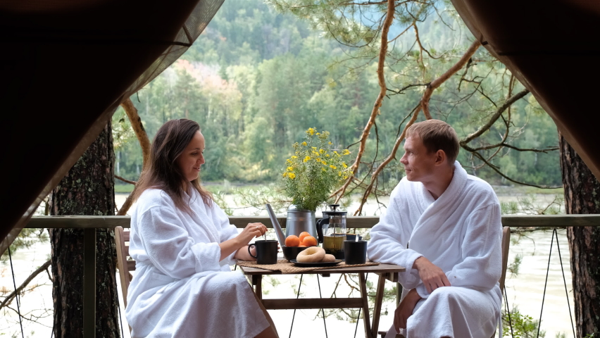 Young Couple in White Terry Bathrobe and Slippers Enjoying Breakfast Outside Glamping Tent. Fast River and Coniferous Forest are on the Background. Luxury Camping, Glamping, Cozy Camping Vacation Royalty-Free Stock Footage #1059045020