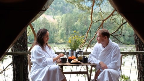 Young Couple in White Terry Bathrobe and Slippers Enjoying Breakfast Outside Glamping Tent. Fast River and Coniferous Forest are on the Background. Luxury Camping, Glamping, Cozy Camping Vacation