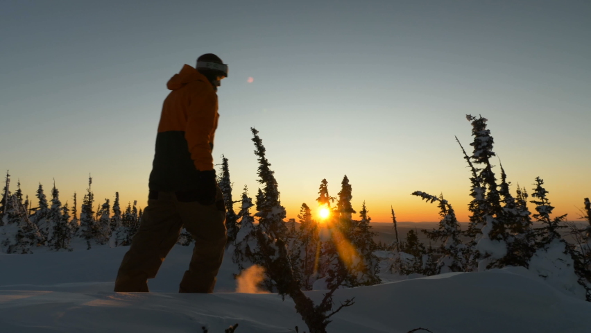 man tourist silhouette wearing warm jacket and gloves walks along snowy valley against fir forest against sunset in cold winter evening
