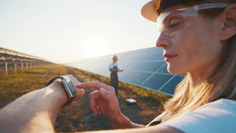 Female smart engineer using smartwatch application software to control solar panel system. Woman technician working at solar farm with modern technologies.