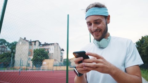 Healthy-looking bearded young athlete after running workout walks on the stadium and uses smartphone fitness application user interface technology.