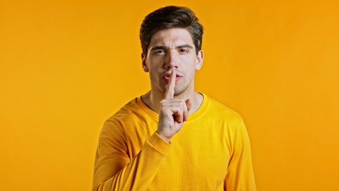 Handsome man holding finger on his lips over yellow background. Gesture of shhh, hush, secret, silence. Close up.
