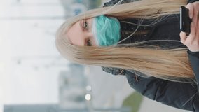 Coronavirus pandemic: blonde woman in a medical mask writing message on smartphone. She walking down the street. Coronavirus pandemic concept. Video with Vertical Screen Orientation 9:16
