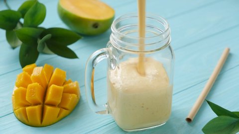Pouring mango yogurt smoothie in glass jar, blue background. Refreshing cold healthy blended drink Mango Lassi