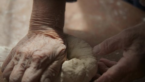Cooking homemade pastries: grandmother kneads soft dough on a wooden table in a room with natural sunlight. The frame contains only wrinkled hands and flour. Home traditions, national cuisine.