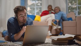 Young man in headset having educational webinar using laptop studying on floor in dorm living room. Diverse girlfriends surfing internet on smartphone relaxing sofa blurred background