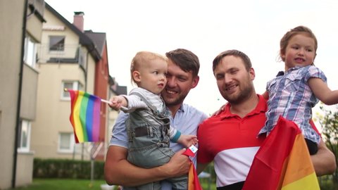 Lovely homosexual couple, two gay men with two adopted children. They hug each other and laugh. Holding rainbow flags, lgbt community