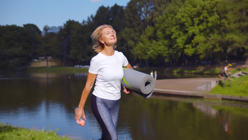 Active senior woman with fitness mat and water bottle in hands walking in park. Happy mature lady strolling in summer park going at outdoors yoga class | Shutterstock HD Video #1059060104