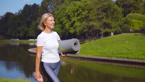 Active senior woman with fitness mat and water bottle in hands walking in park. Happy mature lady strolling in summer park going at outdoors yoga class